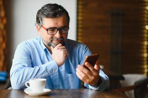 contento empresario sentado en restaurante y esperando para almuerzo. él es utilizando inteligente teléfono y hablando con alguien. negocio personas mayores estilo de vida concepto. foto