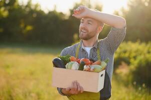 farmer holding a crate of bio vegetables in the farm. Happy man showing box of harvested vegetables. photo