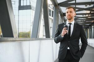 barbado empresario en formal traje en descanso utilizando móvil teléfono utilizar teléfono inteligente negocio hombre en pie fuera de en moderno urbano ciudad calle antecedentes con café taza en céntrico al aire libre. Copiar espacio foto