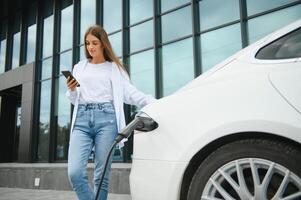 Phone in hands. Woman on the electric cars charge station at daytime. Brand new vehicle. photo