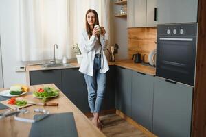 A young beautiful caucasian woman stands in the kitchen with a white cup of coffee or tea in the morning. A lonely girl is enjoying a cup of fresh hot drink. photo