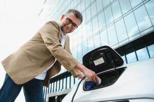 Hansome bearded man sitting near his new modern electric car and holding plug of the charger, while car is charging at the charging station photo