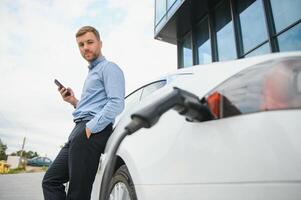 Handsome man in business suit surfing internet on modern smartphone while waiting electric car to charge. photo