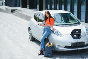 joven mujer es en pie cerca el eléctrico coche. el alquiler coche es cargando a el cargando estación para eléctrico vehículos coche intercambio. foto