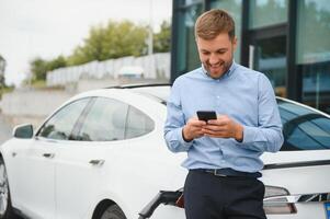 Handsome man in business suit surfing internet on modern smartphone while waiting electric car to charge. photo