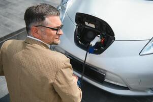 Hansome bearded man sitting near his new modern electric car and holding plug of the charger, while car is charging at the charging station photo