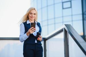 hermosa mujer yendo a trabajo con café caminando cerca oficina edificio. retrato de exitoso negocio mujer participación taza de caliente beber. foto