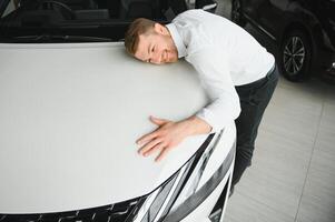 Happy handsome bearded man buying a car in dealership, guy hugging hood of new car photo