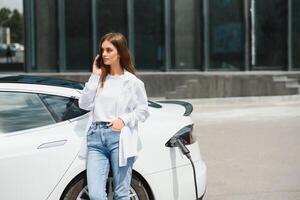 Phone in hands. Woman on the electric cars charge station at daytime. Brand new vehicle. photo