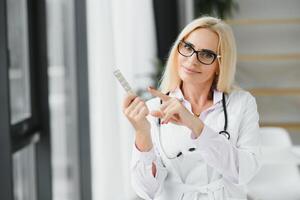 Smiling middle aged female therapist in white uniform holds pills. photo