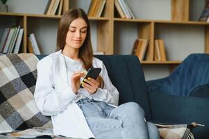 sonriente joven caucásico mujer sentar relajarse en sofá en vivo habitación hablar en vídeo llamar. contento milenario hembra descanso en sofá a hogar tener cámara web digital virtual comunicación en Teléfono móvil. foto