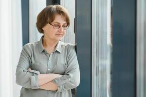 Portrait of beautiful senior woman. Elderly lady is standing by window at home. Active longevity concepts photo