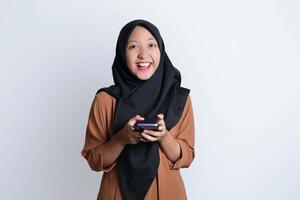 emocionado hermosa asiático mujer en marrón camisa y hijab utilizando móvil teléfono, celebrando éxito, consiguiendo bueno Noticias aislado en blanco antecedentes foto