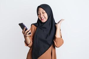 emocionado hermosa asiático mujer en marrón camisa y hijab utilizando móvil teléfono, celebrando éxito, consiguiendo bueno Noticias aislado en blanco antecedentes foto