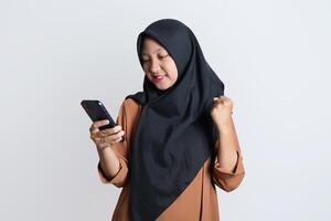 emocionado hermosa asiático mujer en marrón camisa y hijab utilizando móvil teléfono, celebrando éxito, consiguiendo bueno Noticias aislado en blanco antecedentes foto