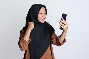 emocionado hermosa asiático mujer en marrón camisa y hijab utilizando móvil teléfono, celebrando éxito, consiguiendo bueno Noticias aislado en blanco antecedentes foto