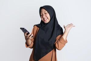 emocionado hermosa asiático mujer en marrón camisa y hijab utilizando móvil teléfono, celebrando éxito, consiguiendo bueno Noticias aislado en blanco antecedentes foto
