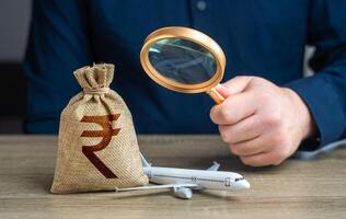 Airplane and indian rupee money bag under investigation. Economic impact of aviation industry. Payment of taxes, fees and excise taxes. Measure impact on local economy. Environmental footprint. photo