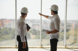 A front view of two smart architects with white helmets reviewing blueprints at a construction site on a bright sunny day photo