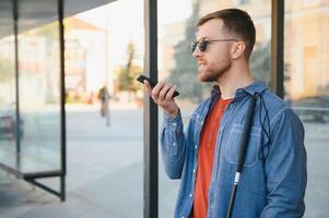 Young blind man with smartphone sitting on bench in park in city, calling. photo