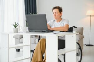 Middle age woman using laptop sitting on wheelchair at home photo