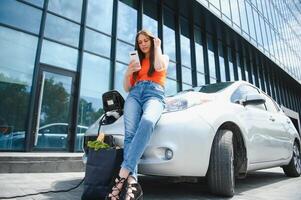 utilizando teléfono inteligente mientras espera. mujer en el eléctrico carros cargar estación a tiempo de día. marca nuevo vehículo. foto