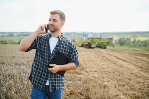 Happy farmer proudly standing in a field. Combine harvester driver going to crop rich wheat harvest. Agronomist wearing flannel shirt, looking at camera on a farmland photo