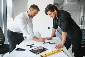 two building designers standing in a modern office leaning over a desk discussing blueprints together photo