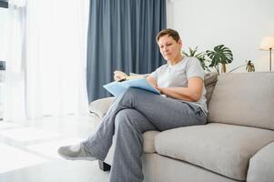 atractivo de edad mediana mujer con un encantador sonrisa sentado en un sofá en el vivo habitación agarrando un libro. foto