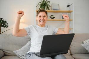 Excited mature woman looking at laptop screen, reading good news in message, celebrating online lottery win, rejoicing success, overjoyed older female sitting on couch at home, using computer. photo