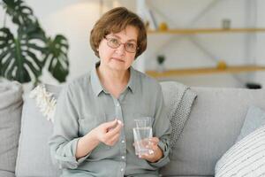 anciano mujer sentado en el sofá y tomando pastillas a hogar. pensionista con medicamentos foto