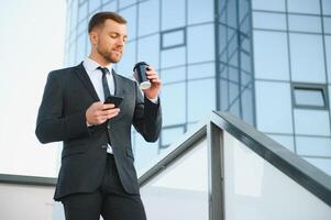 barbado empresario en formal traje en descanso utilizando móvil teléfono utilizar teléfono inteligente negocio hombre en pie fuera de en moderno urbano ciudad calle antecedentes con café taza en céntrico al aire libre. Copiar espacio foto