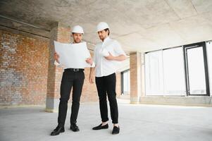 A front view of two smart architects with white helmets reviewing blueprints at a construction site on a bright sunny day photo