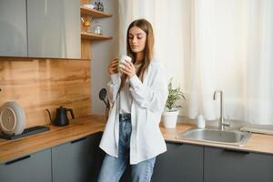 un joven hermosa caucásico mujer soportes en el cocina con un blanco taza de café o té en el Mañana. un solitario niña es disfrutando un taza de Fresco caliente beber. foto