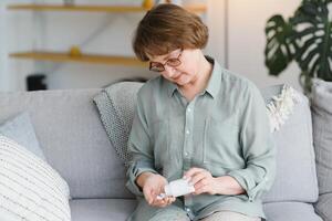 Eldery woman sitting on the sofa and taking pills at home. Pensioner with medicaments photo