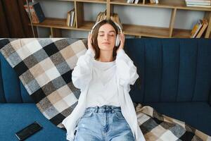 Beautiful young caucasian woman in casual clothing enjoying music and smiling while resting at home. Young woman with headphones using laptop at home photo