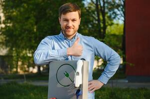 Portrait of a young man standing with charging cable near the charging station. Concept of fast home car chargers photo