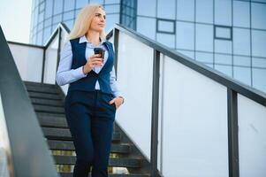 hermosa mujer yendo a trabajo con café caminando cerca oficina edificio. retrato de exitoso negocio mujer participación taza de caliente beber. foto