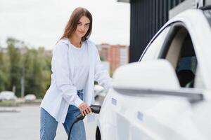 Smiling young caucasian woman plugging electricity cable in electric vehicle for charging on sunny mall parking, cropped. Lifestyle and ecology concept. photo