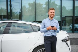 Handsome man in business suit surfing internet on modern smartphone while waiting electric car to charge. photo