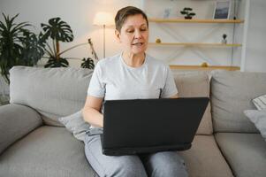 Elderly female sit on couch holding on lap laptop search information, check or typing e-mail, spend time on-line, modern tech, easy interesting pastimes services for retired older generation concept. photo