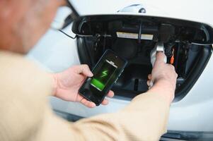 hombre cargando eléctrico vehículo con cable mirando a aplicación en móvil teléfono foto