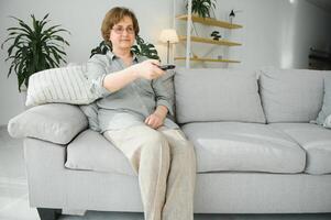 people, television and entertainment concept - senior woman in eyeglasses with remote control watching tv at home photo