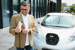 handsome man holding charging cable at electric charging station point standing near his new car. photo