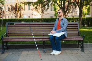 Visually impaired man with walking stick, sitting on bench in city park. Copy space photo