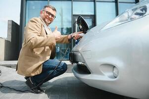 Hansome bearded man sitting near his new modern electric car and holding plug of the charger, while car is charging at the charging station photo