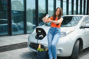 Young woman is standing near the electric car and looks at the smart phone. The rental car is charging at the charging station for electric vehicles. Car sharing. photo