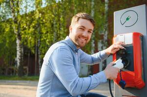 Portrait of a young man standing with charging cable near the charging station. Concept of fast home car chargers photo