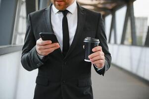 barbado empresario en formal traje en descanso utilizando móvil teléfono utilizar teléfono inteligente negocio hombre en pie fuera de en moderno urbano ciudad calle antecedentes con café taza en céntrico al aire libre. Copiar espacio foto