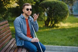 joven ciego hombre con teléfono inteligente sentado en banco en parque en ciudad, vocación. foto